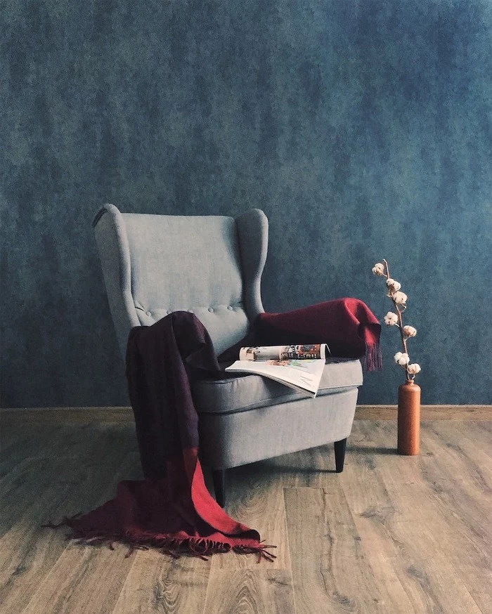wooden floor, grey armchair with throw blanket on it, laminate flooring, blue wall and small vase with flower