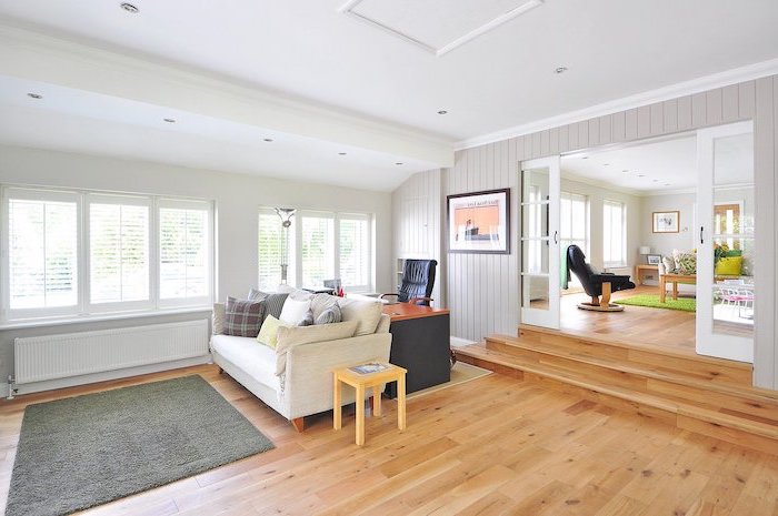 living-room-with-white-sofa-with-throw-pillows-grey-caarpet-on-laminate-flooring-desk-and-chair-white-walls living room with white sofa with throw pillows, laminate flooring, grey carpet on wooden floor, wooden desk with black chair