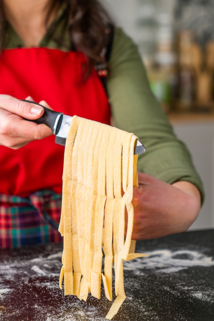long strips of dough, hanging from sharp black knife, tagliatele recipe, black surface covered with flour