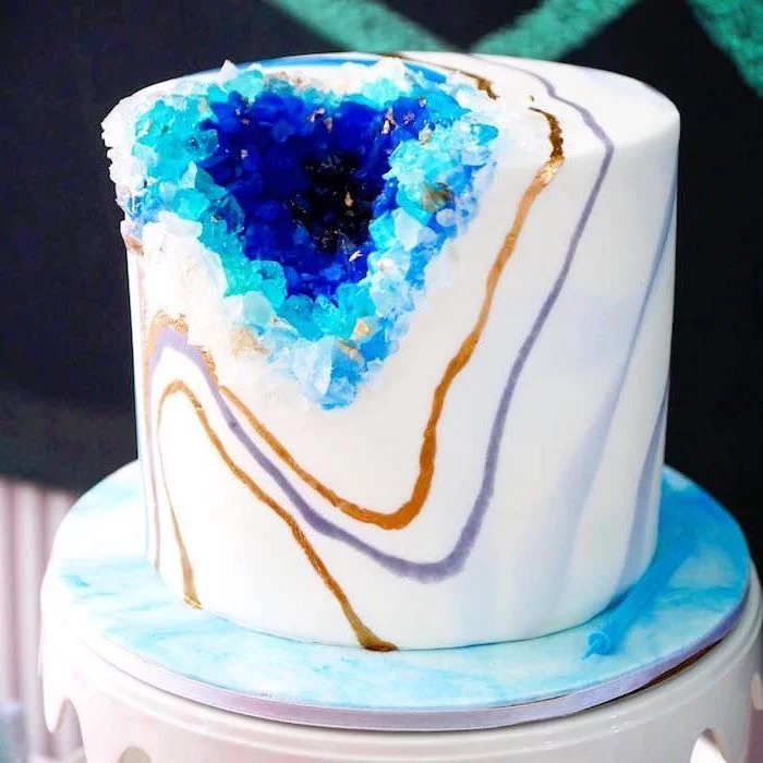 one tier cake, covered with white fondant, decorated with blue rock candy, marble geode cake, placed on white cake stand
