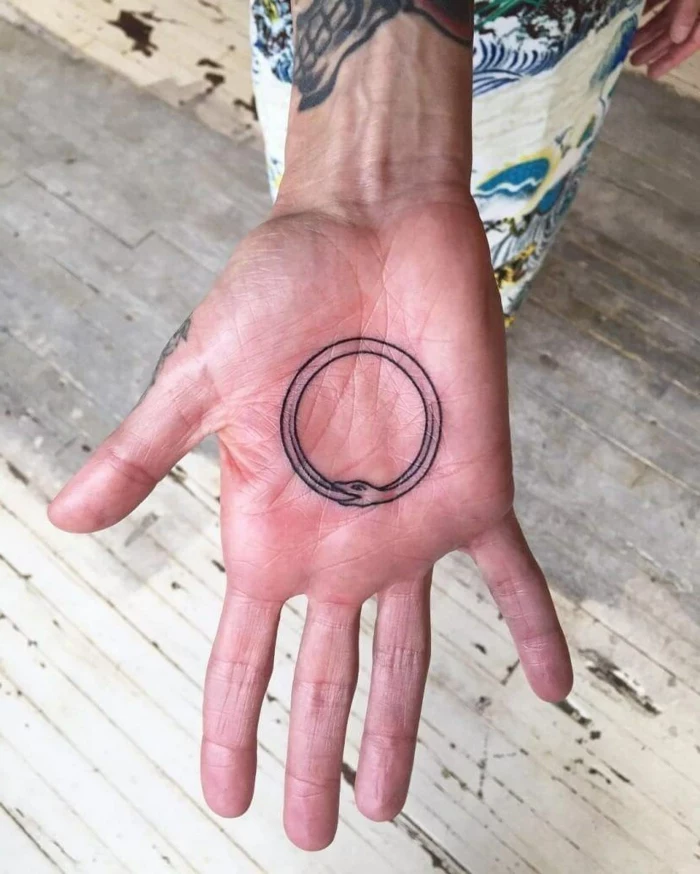 tattoo on the palm of the hand, snake eating its own tail, white wooden floor in the background