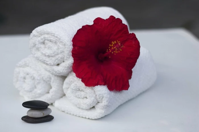 three white towels stacked together, red flower on top of them, spa holidays, three rocks stacked together, all placed on white surface