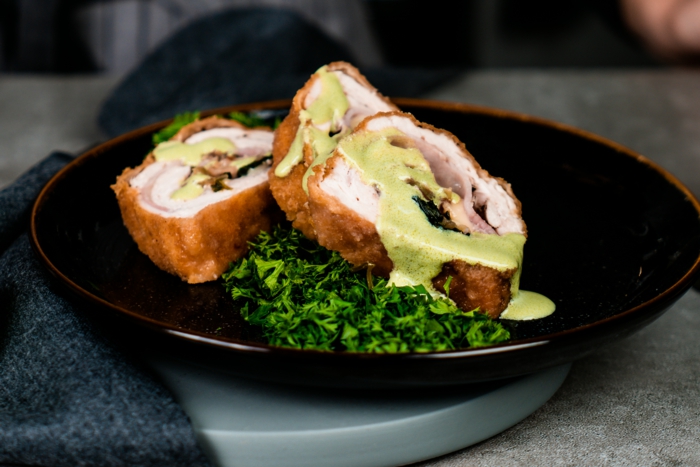chicken cordon bleu recipe, sliced garnished with sauce and kale, placed on black plate