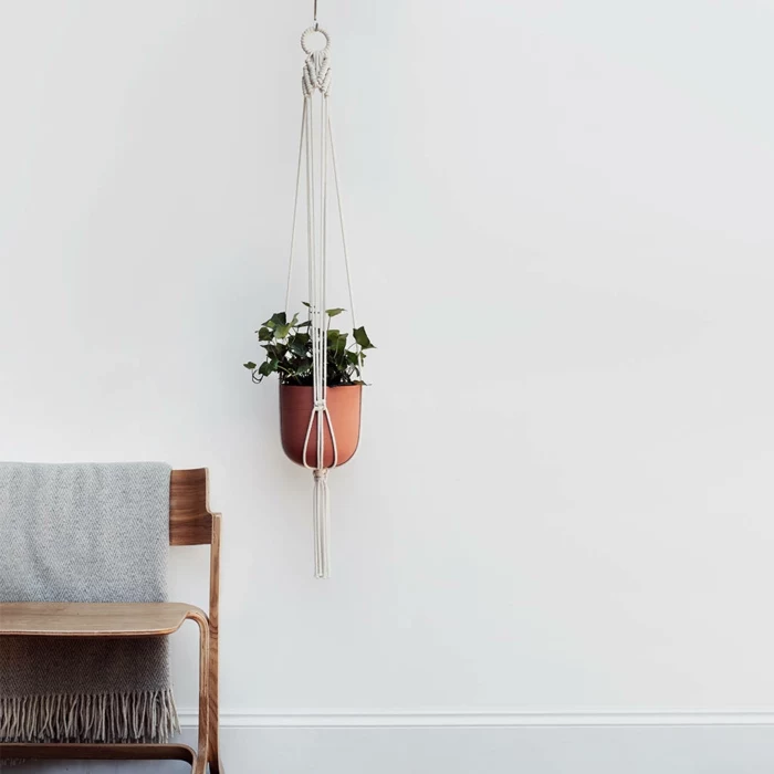 how to make a macrame plant hanger, ceramic pot hanging from the ceiling, next to a wooden chair with throw blanket