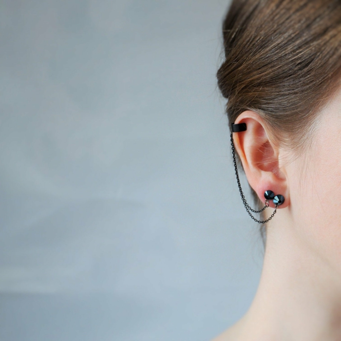 close up photo of an ear, woman with brown hair, cartilage ear piercings, black earrings connected with a chain