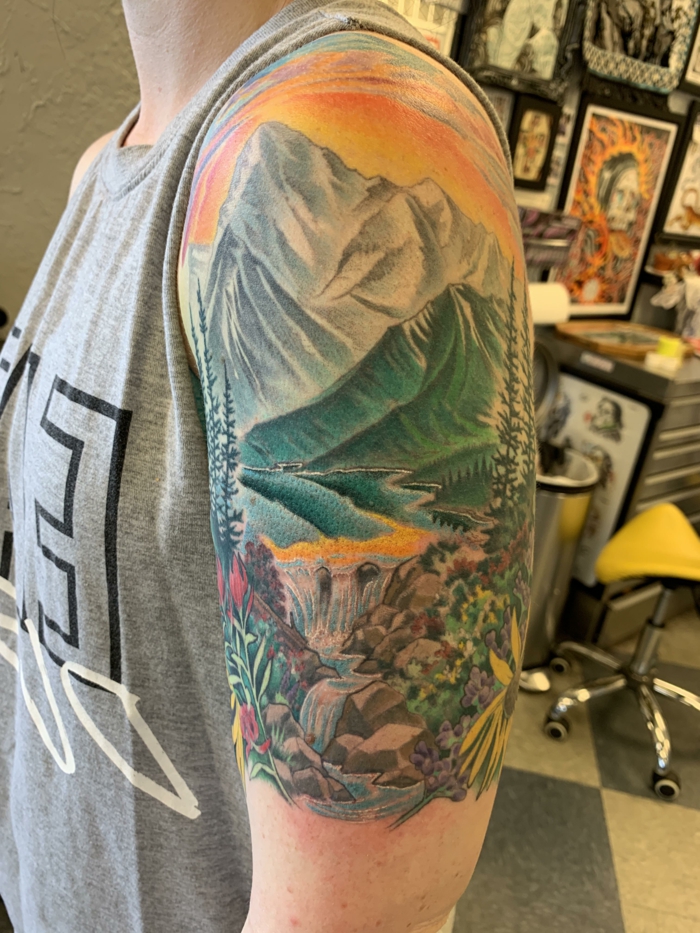 watercolor shoulder tattoo, hiking tattoos, lake surrounded by trees and flowers, mountain range in the background