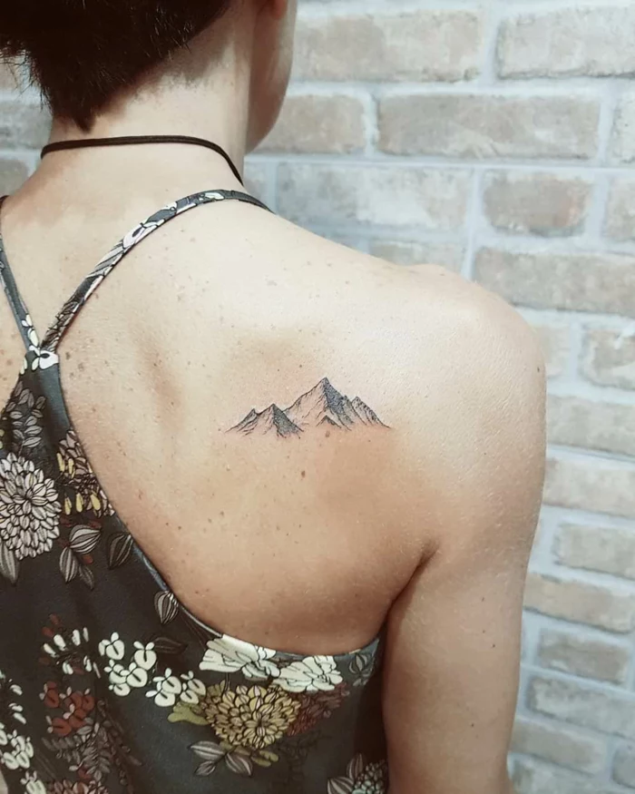 back of shoulder tattoo, minimalist mountain tattoo, woman wearing floral top, brick wall in the background
