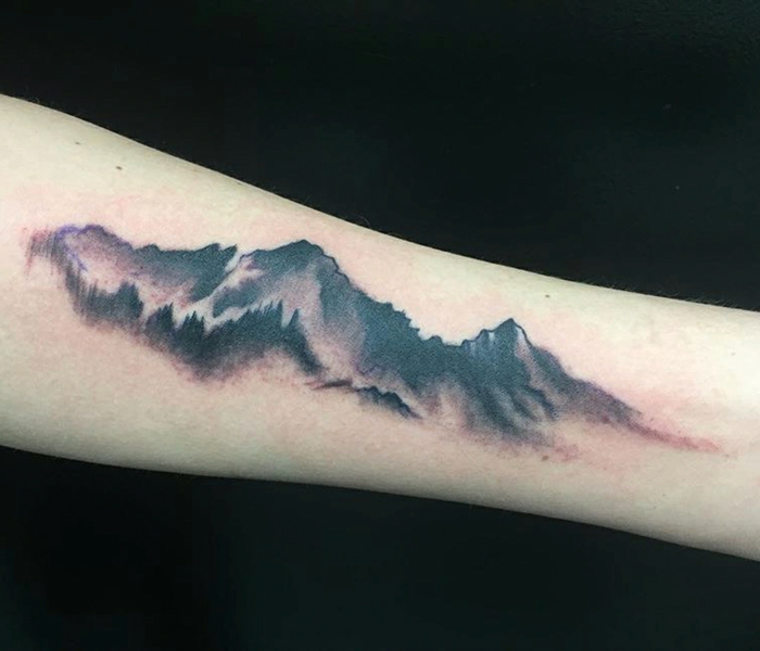 forearm tattoo, mountain tattoo meaning, black background, mountain range with fog and trees