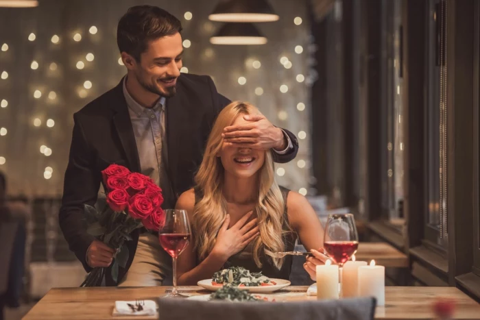 blonde woman sitting at a dinner table, wedding anniversary gifts, man covering her eyes, holding a bouquet of roses