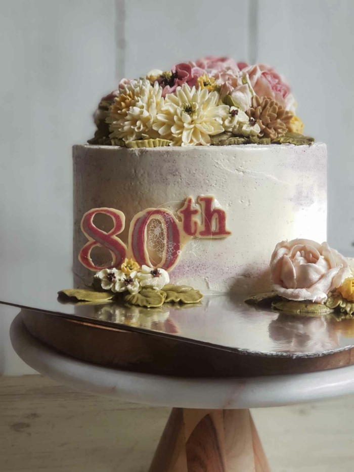 one tier cake covered with white buttercream, decorated with flowers, happy 80th birthday, wooden cake stand