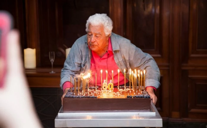 old man standing in front of a table, large chocolate cake on it, blowing out candles, 80th birthday color