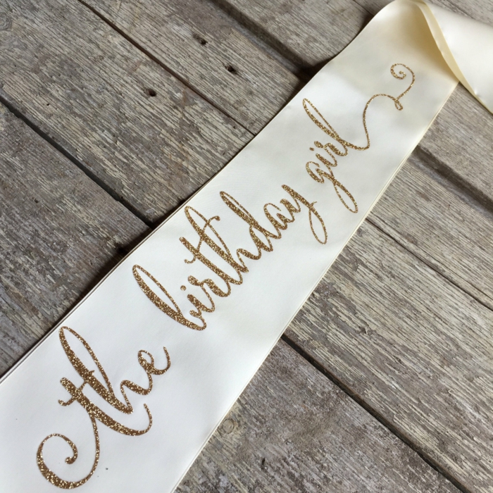 the birthday girl, white sash with gold glitter letters, 18th birthday party ideas, placed on wooden surface