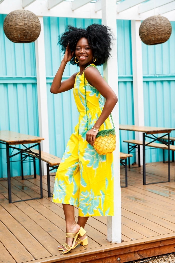 blue wall background cute outfit ideas for summer woman with black curly hair wearing yellow jumpsuit with blue flowers yellow sandals pineapple bag