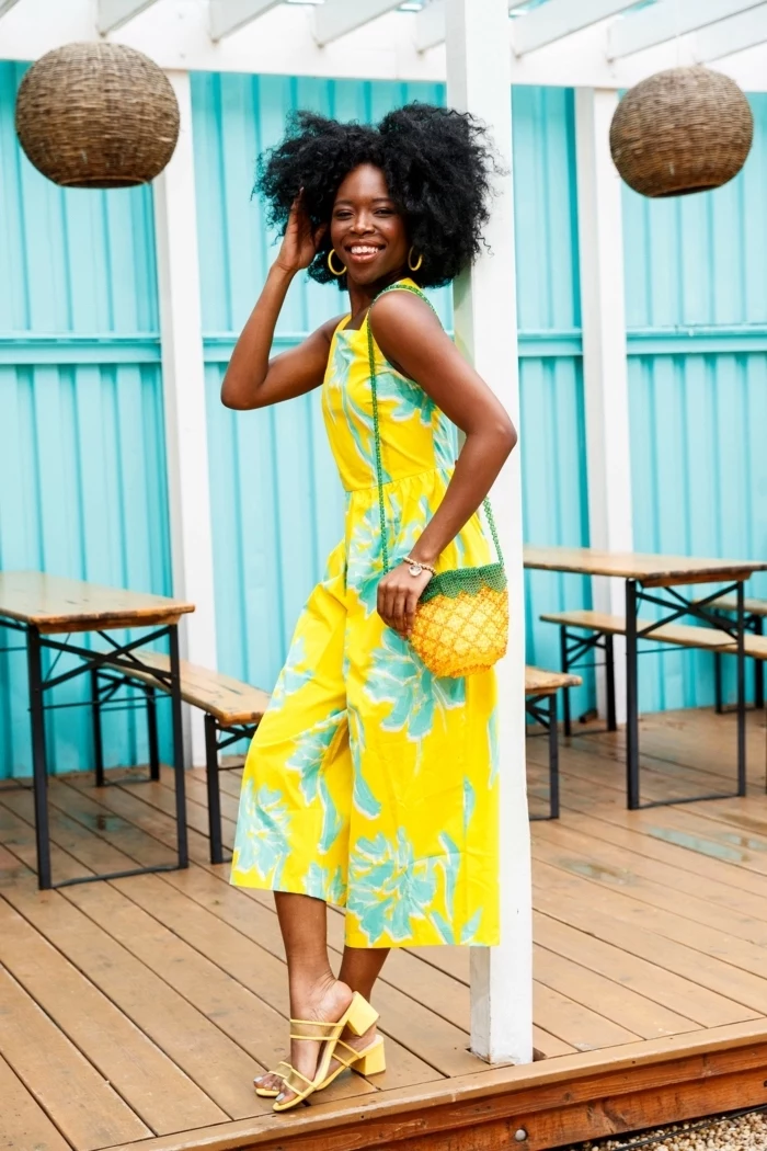blue wall background cute outfit ideas for summer woman with black curly hair wearing yellow jumpsuit with blue flowers yellow sandals pineapple bag
