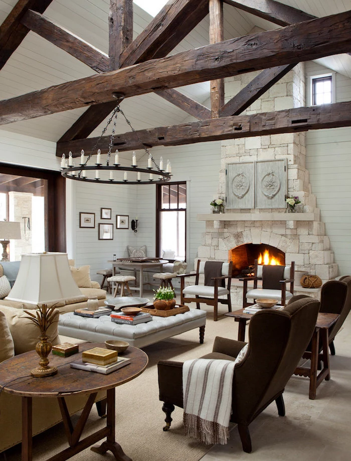 exposed wooden beams on white ceiling, farmhouse decor ideas, stone fireplace, brown velvet armchairs, white sofa and ottoman