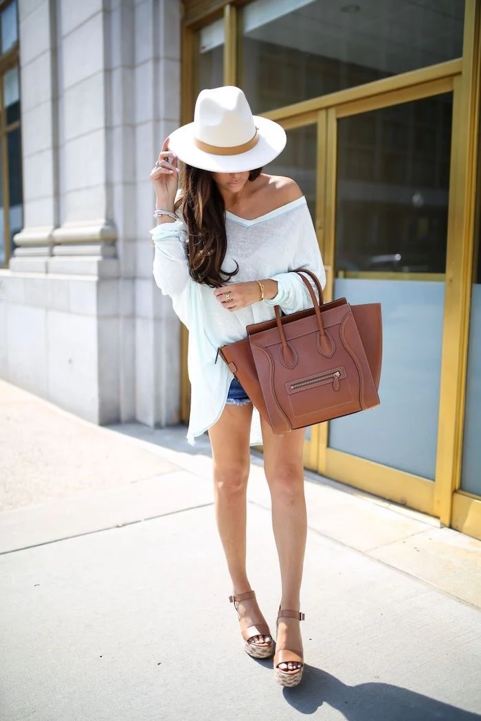 brunette woman wearing white flowy blouse jean shorts white hat cute summer outfits holding brown bag wearing brown leather sandals