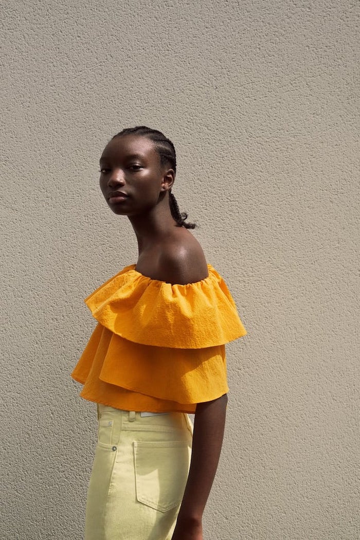 brunette woman with braided hair wearing orange flowy crop top yellow jeans cute outfits for women standing in front of white wall