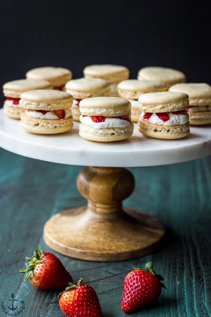 macaroons with white cream and strawberries in the middle summer dessert recipes arranged on wooden cake stand with marble surface