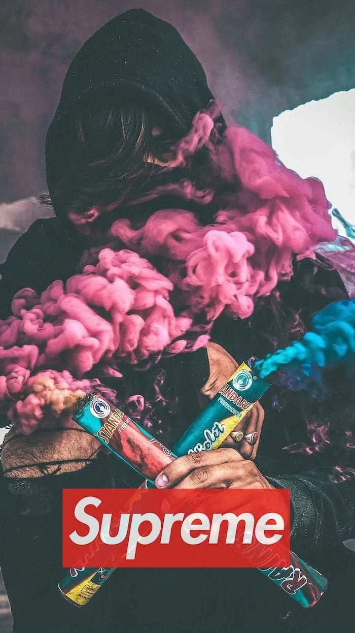 man wearing a hoodie holding to smoke cannons in pink and blue black supreme wallpaper red and white supreme logo