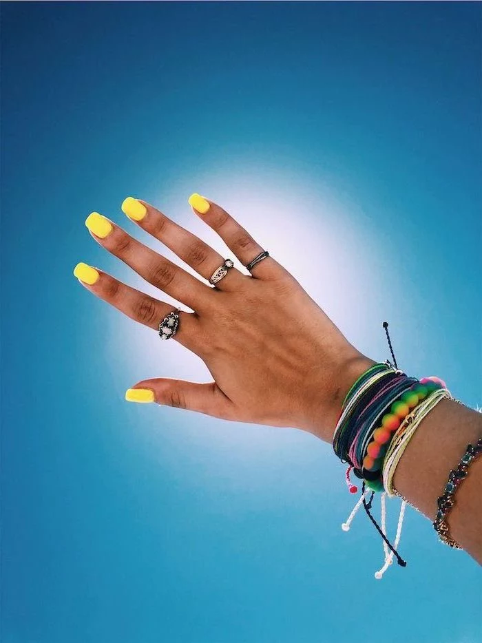 yellow nail polish, medium length square nails, blue background, bracelets on the hand, cute nail ideas