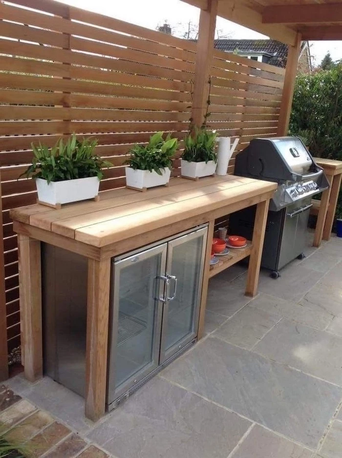outdoor kitchen ideas wooden table with fridge and barbecue grill three potted plants on top stone tiles on the floor