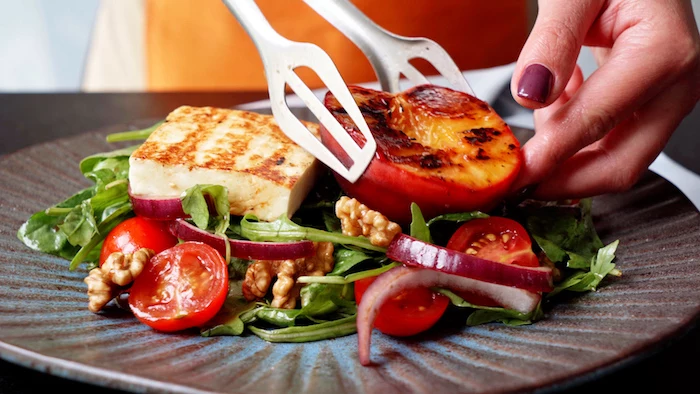 grilled peach and halloumi being placed on top of arugula summer salad recipe with onion cherry tomatoes walnuts