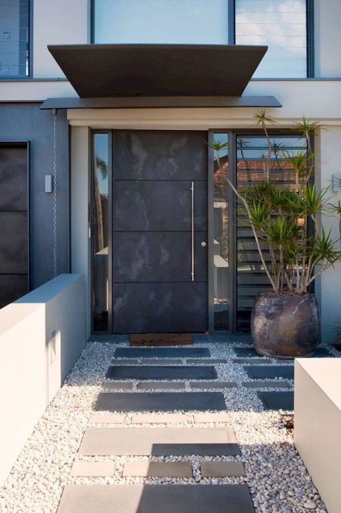 large black door front of house landscaping pot with palm tree next to it tiled gravel pathway