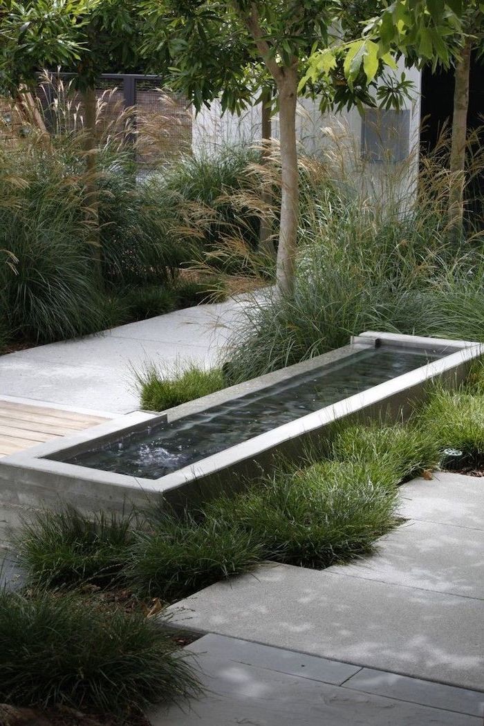 small fountain next to tiled pathway surrounded by bushes and small trees front of house landscaping