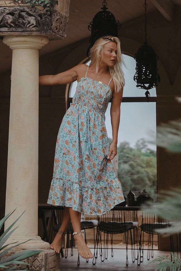 woman with blonde hair wearing midi turquoise dress with orange flowers no shoes standing on a column