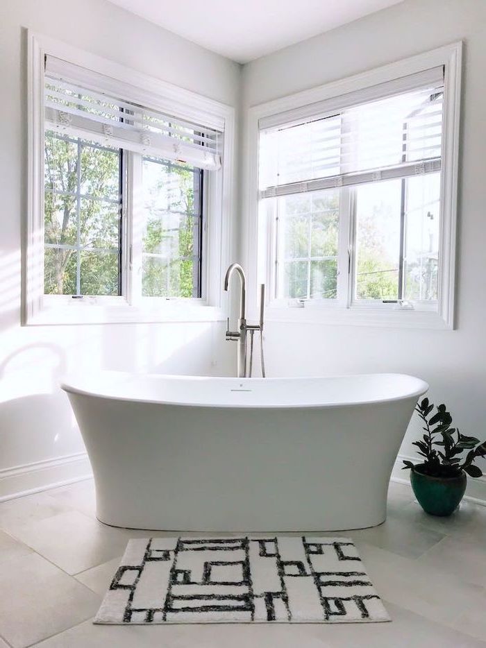 black and white rug in front of white bathtub on white tiled floor white walls with two windows how to decorate a bathroom