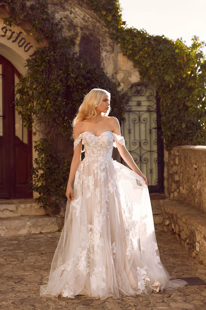 blonde woman with long wavy hair fit and flare wedding dress wearing dress made of tulle and lace standing on paved sidewalk