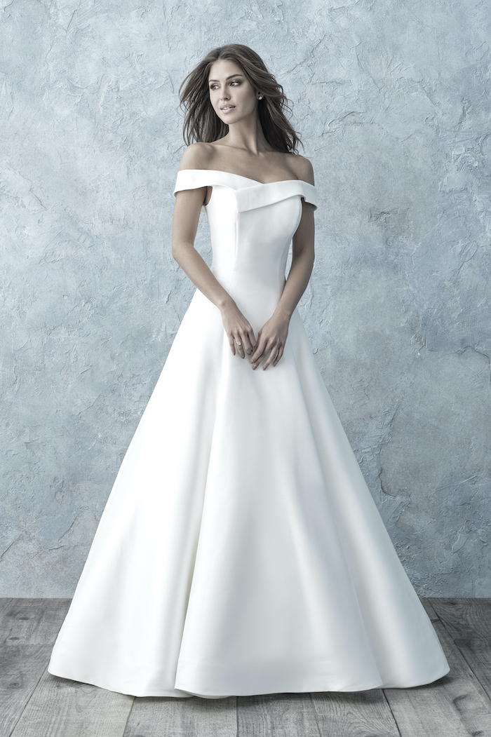 brunette woman wearing long ball gown wedding dress off the shoulders standing on wooden floor in front of blue wall