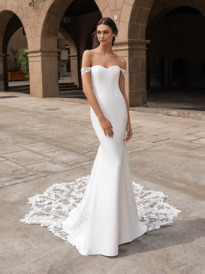 brunette woman wearing long white dress with lace train off the shoulder lace wedding dress standing on sidewalk