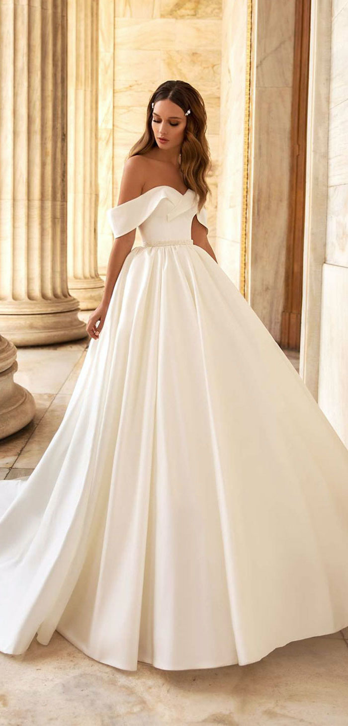 brunette woman with long wavy hair wearing white gown beaded wedding dresses standing in marble hallway