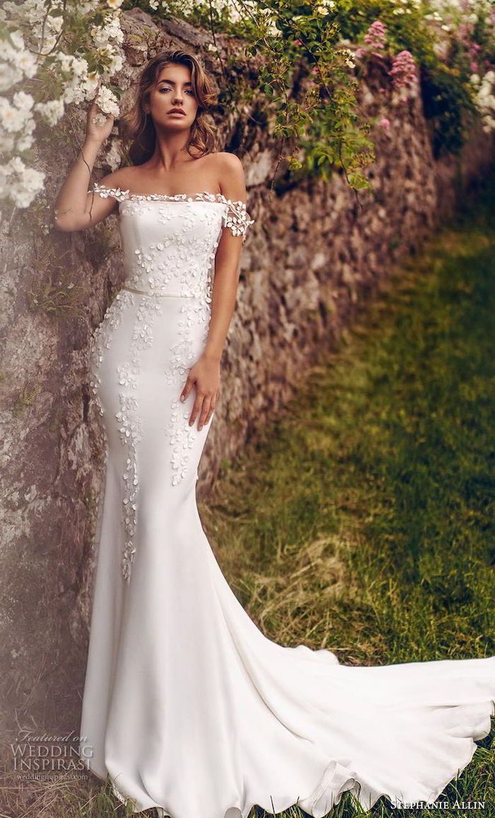 brunette woman with shoulder length hair leaning on stone wall wearing long sleeve lace wedding dress with long train