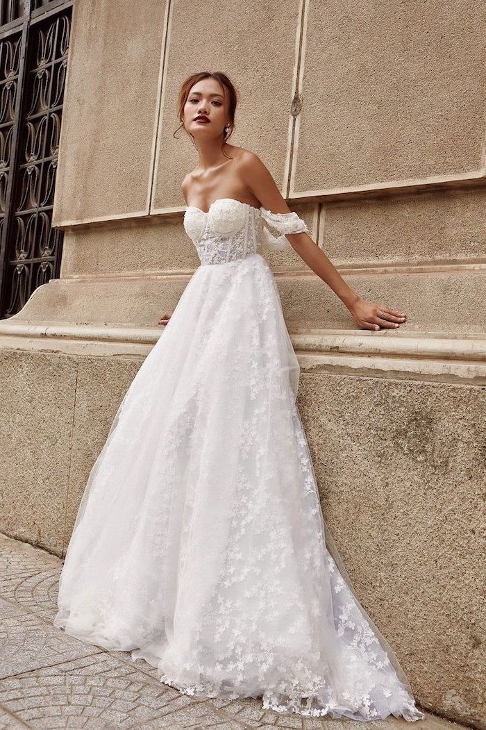 girl with brunette hair in low updo leaning on a wall long sleeve wedding dress wearing white tulle dress with corsette