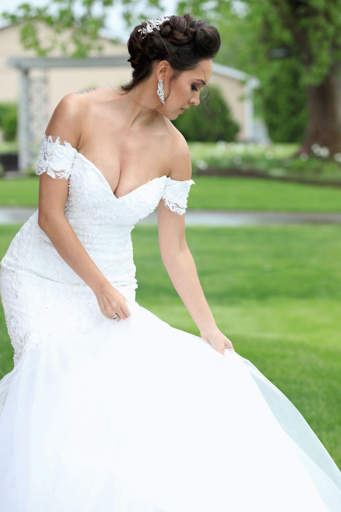 girl with brunette hair in updo wearing a lace and tulle off the shoulder wedding dress standing on green grass field