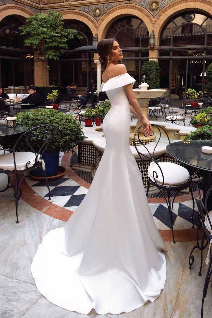 long satin mermaid dress with long train worn by brunette woman fit and flare wedding dress standing in an outside restaurant