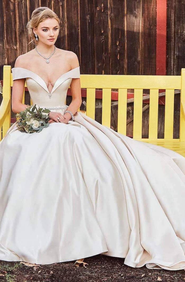 long sleeve wedding dress blonde woman sitting on yellow bench holding a bouquet wearing long satin dress with train