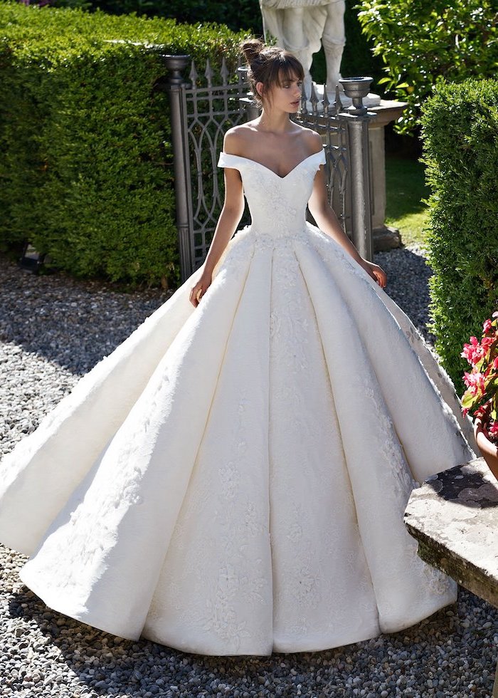 off the shoulder long sleeve wedding dress brunette woman with high updo and bangs wearing white gown on gravel pathway