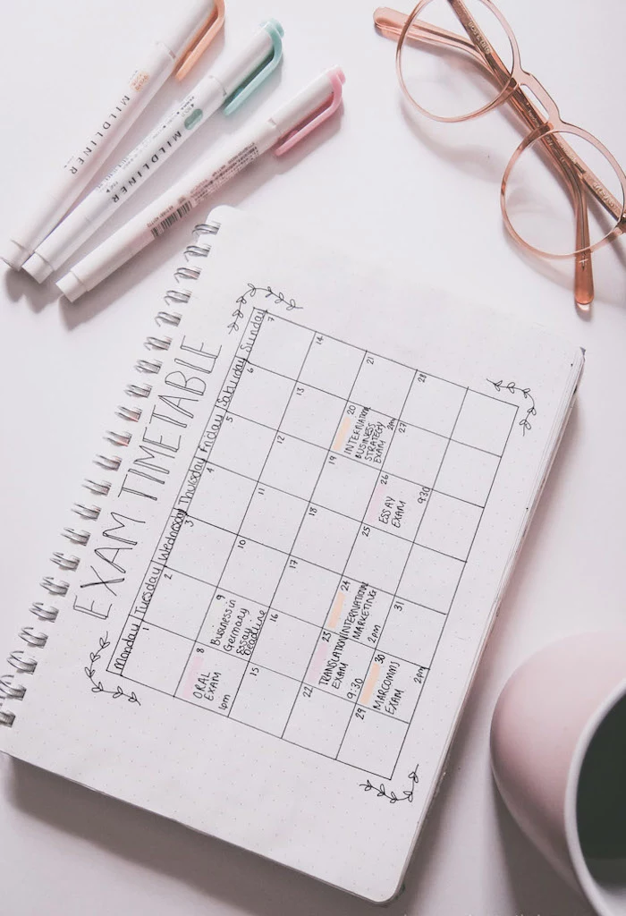 what is a bullet journal exam timetable schedule written on white notebook coffee cup pens and glasses on white surface next to it