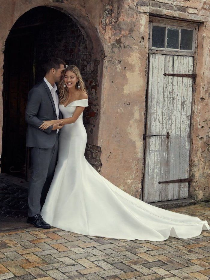 woman and man standing on paved sidewalk off the shoulder wedding dress wearing suit and white dress with long train