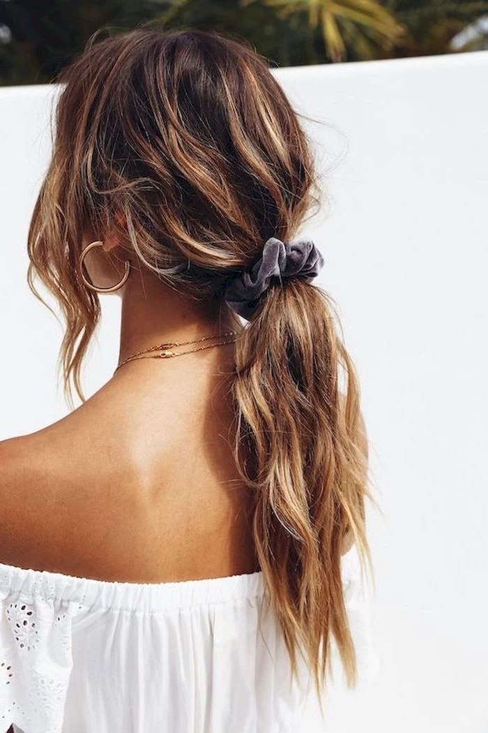 woman wearing strapless white top easy hairstyles for long hair brunette wavy hair with blonde highlights in low ponytail tied with velvet scrunchie