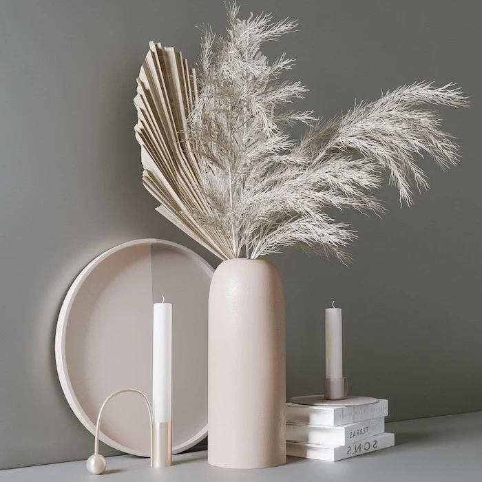 arrangement with white faux pampas grass two candles placed on top of three books with white covers gray wall