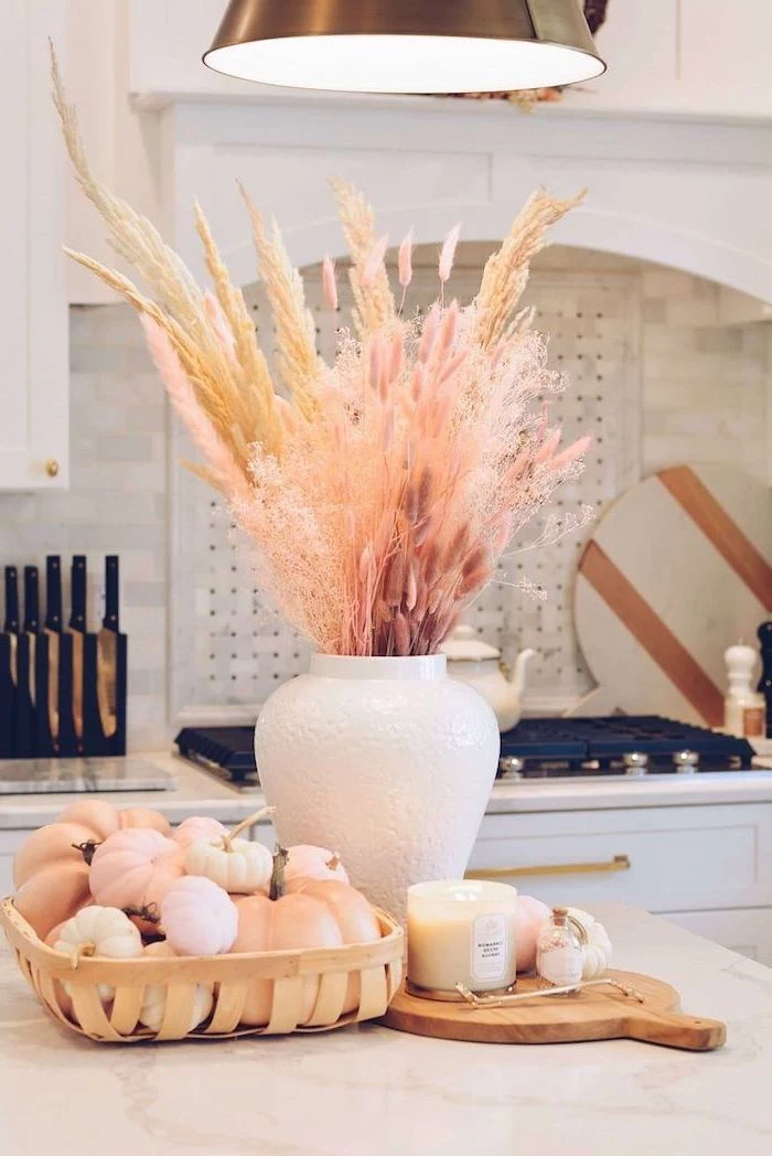 artificial pampas grass inside white ceramic vase placed on top of kitchen island wooden basket with small pumpkins inside