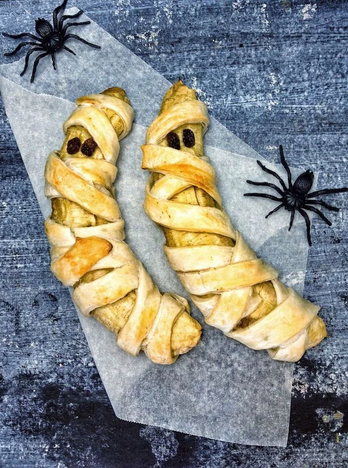 bananas wrapped in dough as mummies placed on baking paper halloween themed food two plastic spiders on the sides