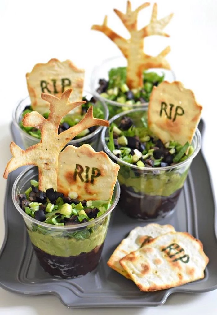 black beans and guacamole dip garnished with chives chopped olives inside small cups halloween themed food crackers with rip written on them