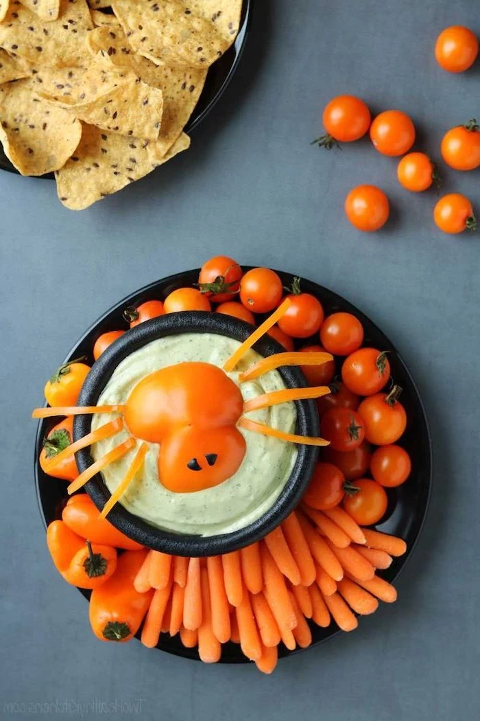 black plate with cherry tomatoes baby carrots orange peppers arranged around bowl of guacamole dip halloween snack ideas placed on gray surface