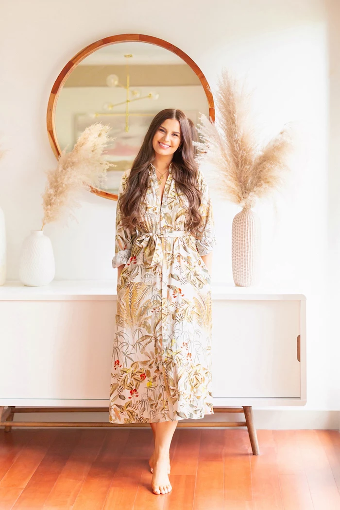brunette woman wearing colorful dress leaning on white cupboard with two ceramic vases with colored pampas grass round mirror on white wall