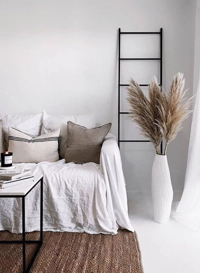 dried pampas grass inside tall white vase standing next to sofa covered with white blanket throw pillows white walls and floor